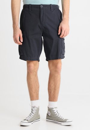 Homme portant un short cargo noir, des baskets montantes grises avec des lacets blancs et des chaussettes blanches, debout devant un fond clair uni.