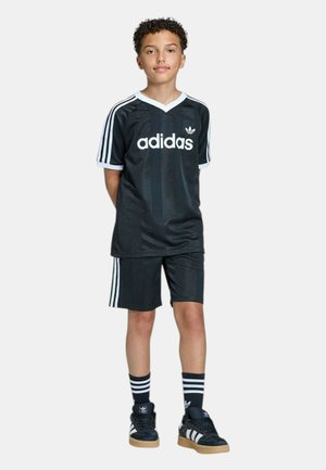 Ragazzo in piedi con le mani dietro la schiena, indossa abbigliamento sportivo nero Adidas, calzini a righe e scarpe da ginnastica Adidas nere su sfondo uniforme.