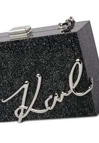 Melna spīgākā klučs, izgatavots no cieta materiāla, ar metāla aizdari un ķēdi. Priekšpusē ir sudraba slīpas rakstības "Karl" dizains.