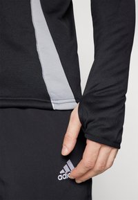 T-shirt de sport noir à manches longues avec des panneaux latéraux gris. Il dispose d'ouvertures pour les pouces dans les manches et d'un logo Adidas en bas.