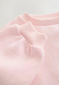 Tela de sudadera en rosa claro con cuello y puños acanalados, que presenta una textura suave y superficie lisa. El diseño incluye detalles recogidos en las mangas.