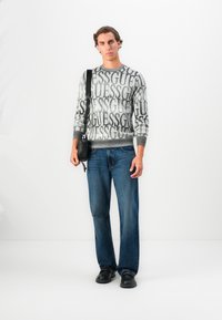 Uomo che indossa un maglione grigio con il testo "GUESS", jeans blu, scarpe nere e una borsa a tracolla nera, in piedi di fronte a uno sfondo bianco.