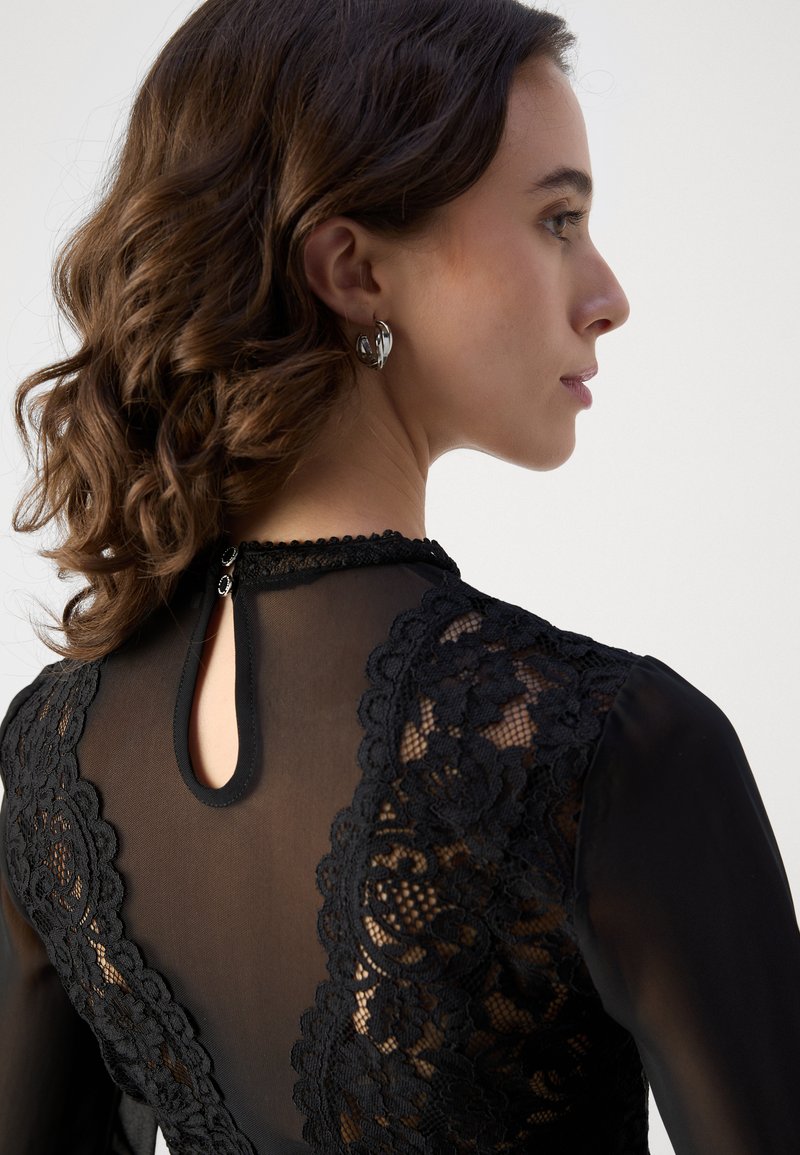 Blusa negra con detalles de encaje en la parte posterior, tejido transparente, escote redondo y un corte en forma de ojal con cierre de botón.