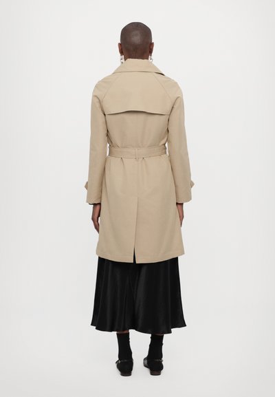 Beige trench coat with a belted waist, back vent, and flap detailing. Worn over a black satin skirt and black footwear.