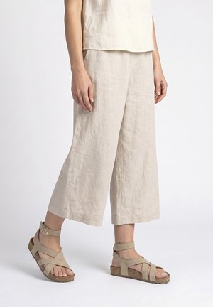 Persoon draagt beige, wijde cropped broek en beige sandalen met bandjes en kurken zolen, staand tegen een witte achtergrond.