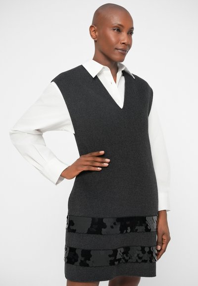 Vestido sem mangas de lã cinza com decote em V, adornado com duas faixas de lantejoulas pretas na bainha, combinado com uma camisa branca com colarinho por baixo.