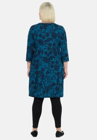 Robe teal à mi-longueur avec un motif floral sombre, manches trois-quarts et design évasé, portée avec des leggings noirs et des ballerines.