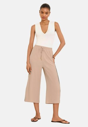 Frau trägt ein ärmelloses weißes V-Ausschnitt-Top, beige weite, kurze Hosen mit Kordelzug und dunkle flache Sandalen, steht mit einer Hand an der Hüfte.