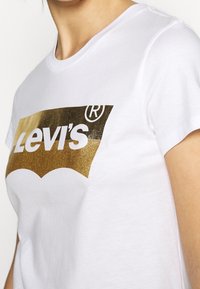 Biały bawełniany T-shirt z dużym złotym brokatowym logo Levi's i łukowatym wzorem. Krótkie rękawy i okrągły dekolt.