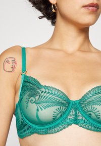 Sutiã de renda teal com intrincada bordadura espiral, copos de malha transparente, alças ajustáveis e acabamentos com recortes em forma de ondas. Detalhes em tom dourado.