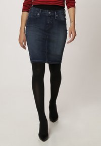 Jeanspencilkjol i mörkblått, med en figurnära silhuett, fem fickor och ljus blekning. Matchad med svarta strumpbyxor och spetsiga klackar.