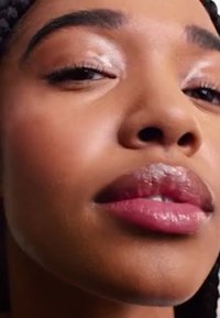 Nærbillede af et ansigt, glat hud, fyldige læber med glansfinish, mørke øjenbryn og subtil øjenmakeup mod en neutral baggrund.