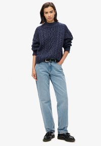 Suéter de punto trenzado azul marino con cuello alto, combinado con jeans sueltos de un azul claro y zapatos negros. Cinturón simple como acento.