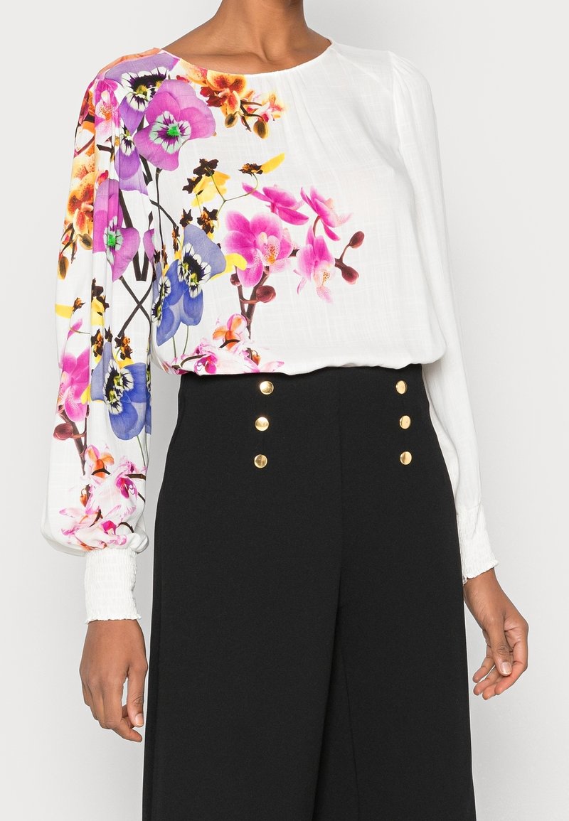 Femme portant un chemisier blanc à manches longues avec un imprimé floral coloré sur le côté gauche et un pantalon noir taille haute avec des boutons dorés.