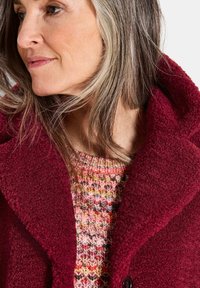 Frau mit schulterlangem grauem Haar, die einen mehrfarbigen Strickpullover und einen strukturierten tiefroten Mantel mit breiten Revers trägt.