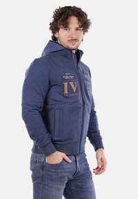 Felpa con zip color navy in materiale morbido, caratterizzata da dettagli ricamati, tasche a marsupio e polsini a costine con una vestibilità aderente.