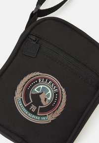 Czarna nylonowa saszetka na zamek błyskawiczny z okrągłą naszywką z haftem przedstawiającym góry i ozdobami laurowymi, oznaczona "ELLESSE CERVINIA ALPINE SKI 78."