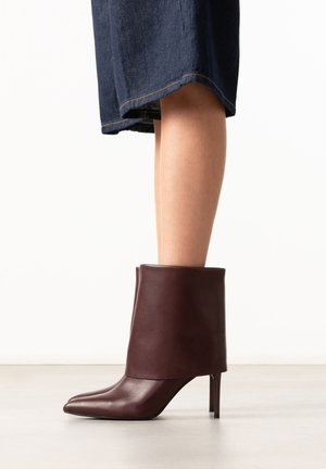 Vrouw draagt donkerbruine leren stiletto laarzen met spitse neus tot halverwege de kuit en een omgeslagen boord, gecombineerd met een donkerblauwe denimrok.