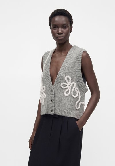 Mannequin à la peau foncée porte un gilet sans manches gris tricoté avec une broderie florale blanche et un pantalon noir taille haute, posant devant un fond blanc.