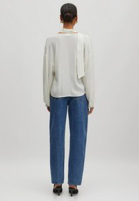 Off-White Bluse mit langen Ärmeln und einem gebundenen Detail am Rücken, kombiniert mit high-waisted blauen Jeans. Enge Passform, glatte Textur und minimale Akzente.
