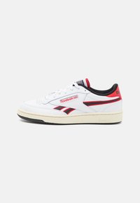 Baskets basses blanches Reebok avec des accents rouges et noirs, semelle beige et doublure intérieure noire, vues de côté sur un fond blanc.