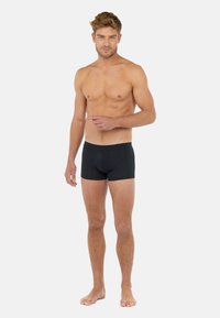 Svarta bomullsboxershorts med en åtsittande design, slät textur och en logobeklädd midja, kombinerat med barfotad och en neutral bakgrund.