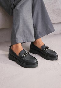 Zwarte leren loafers met een chunky zool en zilveren kettingdetails aan de voorkant. Draag ze met grijze wijde broekspijpen. Duurzaam, gestructureerd oppervlak.