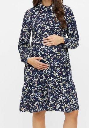 Gravid kvinde iført en langærmet navy blomstret kjole, hænderne blidt støttende hendes mave mod en ensartet hvid baggrund.