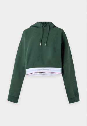 Jeune femme portant un sweat à capuche oversize vert foncé et un pantalon large assorti, debout devant un fond clair uni.