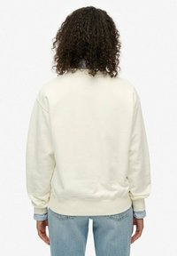 Sudadera crema clara con cuello redondo, puños y dobladillo de canalé, combinada con jeans azul claro, dejando ver una camisa a rayas debajo.
