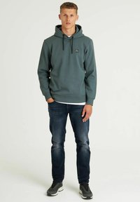 Groene hoodie met trekkoord, voorzak en logo-accent, gecombineerd met donkere spijkerbroek en grijze sport schoenen, staand voor een witte achtergrond.