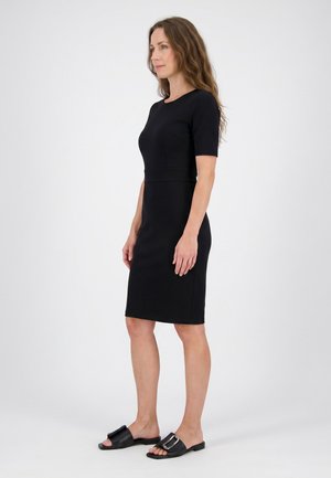 Frau mit langen, welligen Haaren, die ein knielanges schwarzes Kleid mit kurzen Ärmeln und schwarze Slip-on-Sandalen mit Schnallen trägt, steht vor weißem Hintergrund.