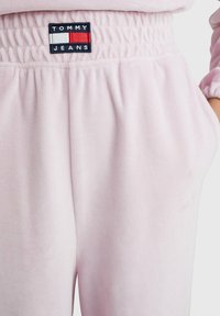 Rožinės veliūro sportinės kelnės su susitraukusia juosta. Juosmens juostoje įsiuvinėtas Tommy Jeans logotipas mėlynos, raudonos ir baltos spalvų.
