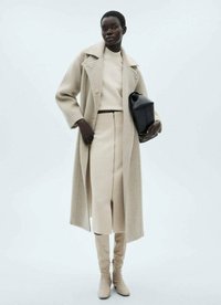 Beige Wollmantel über einem passenden Strickoberteil und -rock mit Reißverschlussdetails. Beige kniehohe Stiefel und eine strukturierte schwarze Handtasche runden den Look ab.