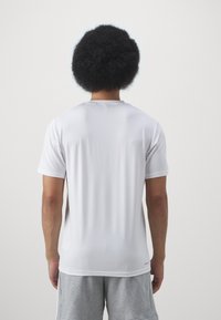T-shirt de sport blanc à manches courtes avec un design sobre et une texture lisse. Il présente un col rond et un logo subtil en bas à gauche.