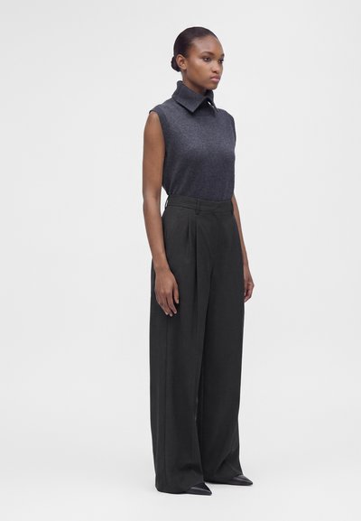 Femme portant un pull sans manches gris foncé à col roulé et un pantalon noir taille haute à jambes larges, debout devant un fond blanc.