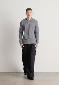 Graues langärmliges Poloshirt mit Kragen, kombiniert mit lockeren schwarzen Cargo-Hosen. Das Shirt zeigt ein dezentes Logo und eine glatte Stofftextur.