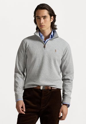 Man in een lichtgrijze kwart-rits trui over een blauwe shirt, donkere bruine broek, een bruine riem en een polshorloge, staande tegen een witte achtergrond.