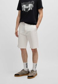 Shorts en coton blancs avec poches latérales, associés à un t-shirt noir avec un imprimé graphique, des baskets grises et des chaussettes blanches avec un logo.