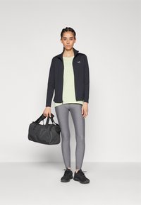 Chaqueta negra con cremallera sobre una camiseta verde clara, leggings grises y zapatillas negras. Sosteniendo un bolso de deporte negro. Texturas suaves y ajuste atlético.