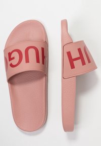 Růžové gumové klouzavé sandály s texturem na popruhu, na kterém je nápis "HUGO" tučnými červenými písmeny. Hladká podrážka, tvarovaný design pro pohodlí.