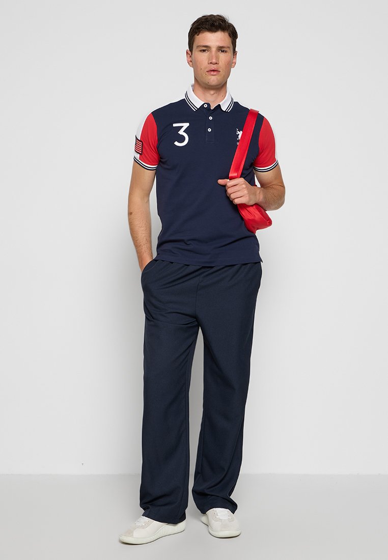 Polo shirt navy avec des manches courtes rouges et des rayures blanches, ornée d'un grand "3" et d'un logo. Associé à un pantalon foncé et des baskets blanches.