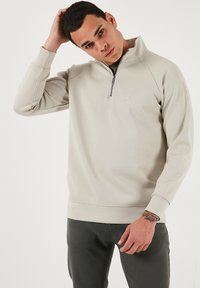 Svijetlo siva dukserica na zip s visokim ovratnikom, raglan rukavima i malim logom. Mekana textura te rebrasti rukavi i donji rub.