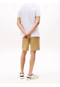 Chemise blanche à manches courtes et shorts kaki, figure debout avec les mains jointes derrière le dos, portant des baskets blanches avec des accents noirs.