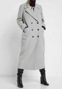 Szary dwurzędowy płaszcz z mieszanki wełny z wciętym kołnierzem, dużymi czarnymi guzikami, bocznymi kieszeniami i długimi rękawami; noszony na czarnej bluzce.