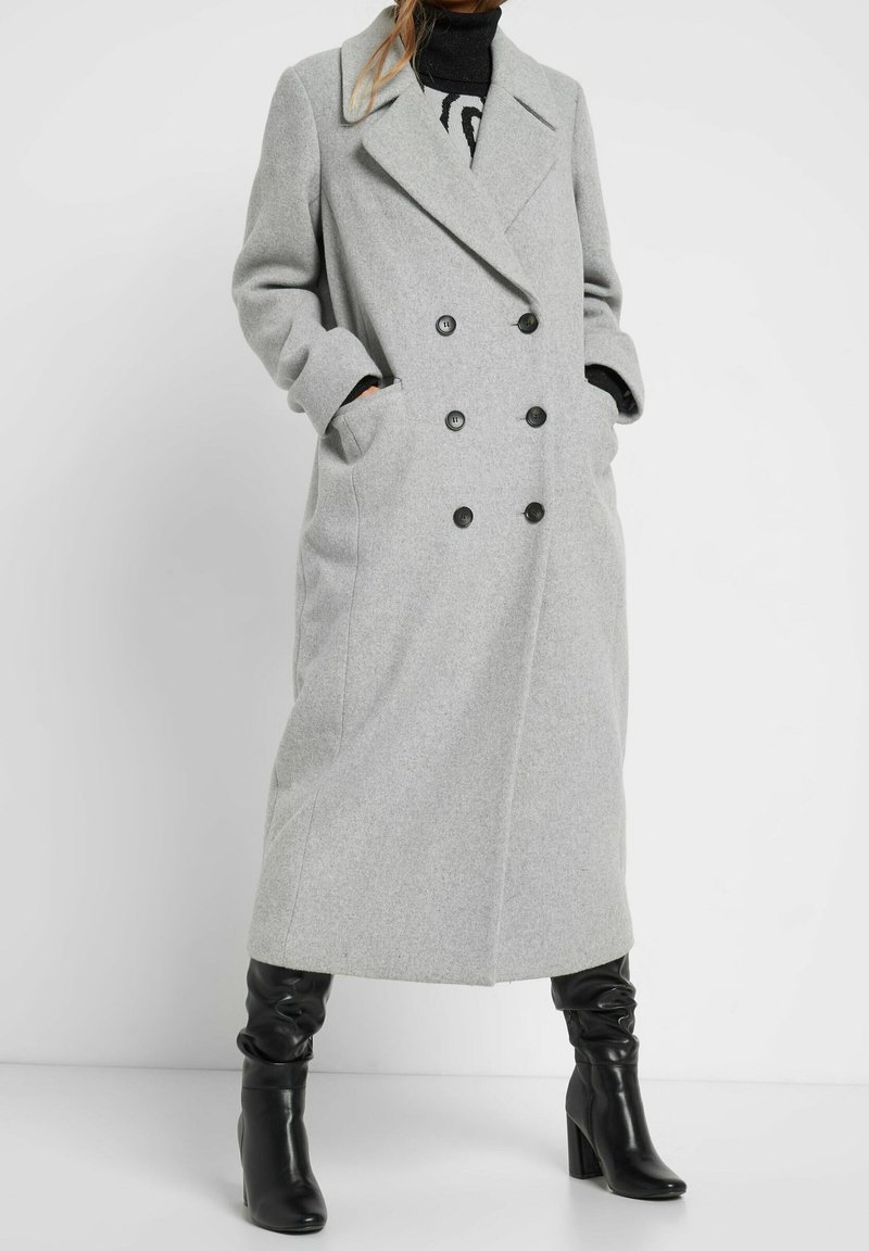 Szary dwurzędowy płaszcz z mieszanki wełny z wciętym kołnierzem, dużymi czarnymi guzikami, bocznymi kieszeniami i długimi rękawami; noszony na czarnej bluzce.