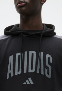 Μαύρο φούτερ με κουκούλα από βαμβακερή σύνθεση, με μεγάλο γκρι λογότυπο «ADIDAS» και ριγωτές λεπτομέρειες στους ώμους. Διαθέτει κορδόνια στην κουκούλα.