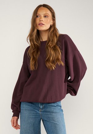 Donna con lunghi capelli castani che indossa una felpa larga viola scuro e jeans azzurri, in piedi davanti a uno sfondo chiaro e semplice.