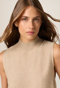 Suéter sin mangas beige con un alto cuello acanalado, tejido texturizado y sutiles detalles brillantes. Superficie suave con un diseño ajustado.