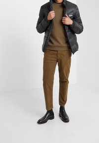 Mann trägt eine schwarze Steppjacke, einen olivgrünen Pullover, braune Hosen und schwarze Leder-Chelsea-Boots vor einem schlichten weißen Hintergrund.